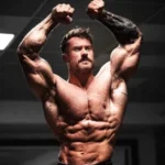 Who is Chris Bumstead - Six-time Mr. Olympia Classic Physique champion posing on stage with trophy and gold medal.