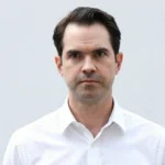 Jimmy Carr in a sharp charcoal suit leans against a black Rolls-Royce, with the London skyline behind him, representing his £25 million Jimmy Carr net worth and comedy career.