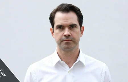 Jimmy Carr in a sharp charcoal suit leans against a black Rolls-Royce, with the London skyline behind him, representing his £25 million Jimmy Carr net worth and comedy career.